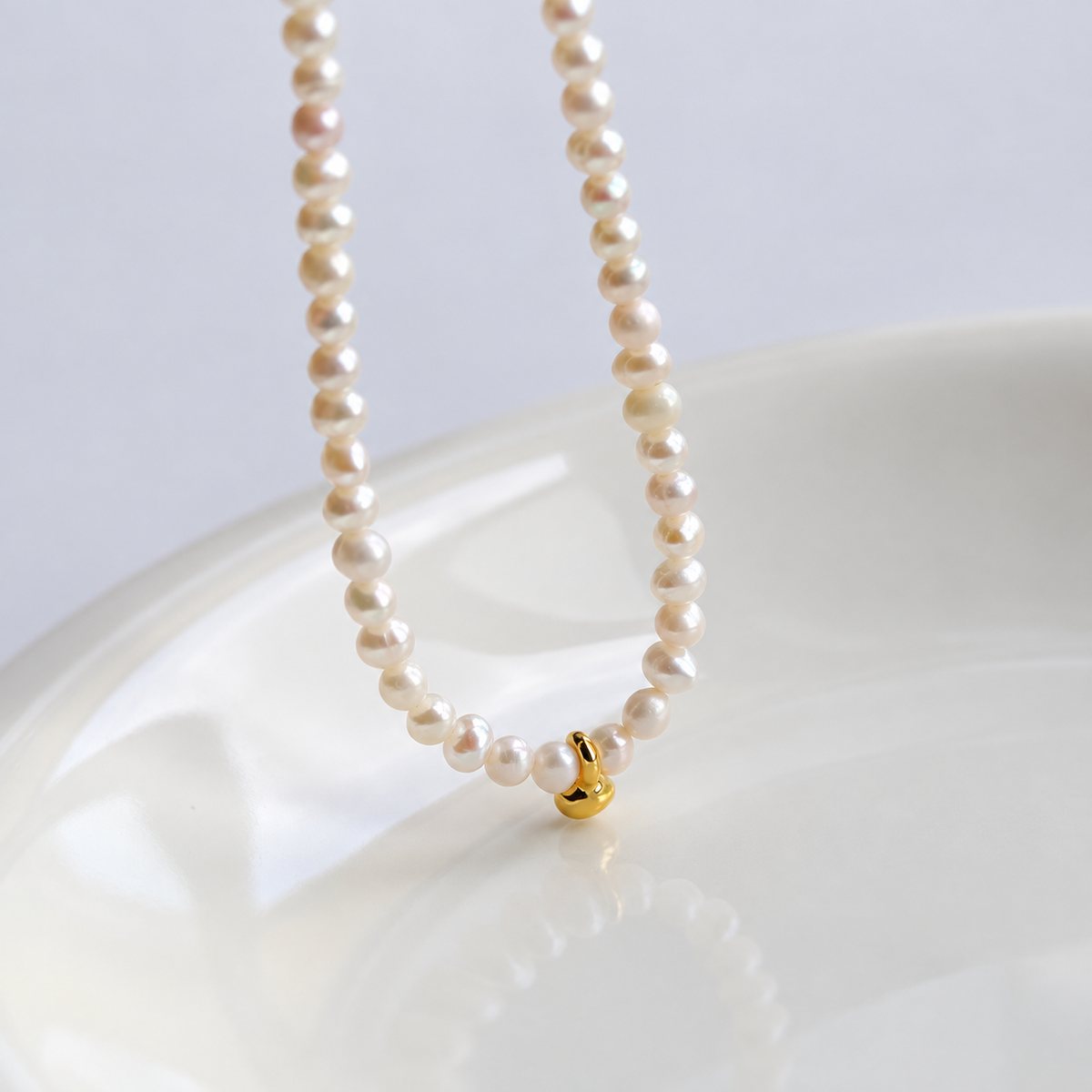 Close-up of a white pearl necklace with a small curved gold-tone bean pendant above an ivory ceramic dish.